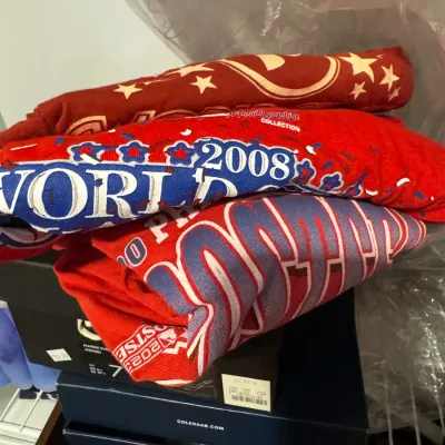 Sale Photo Thumbnail #48: Stack of red sports t-shirts, including a 2008 World Series shirt and a 2023 postseason shirt, resting on shoe boxes.