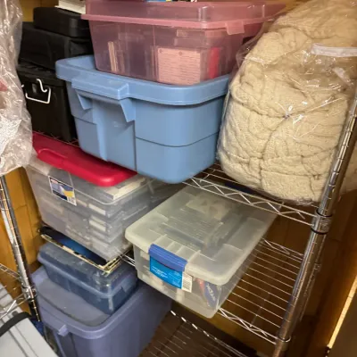 Sale Photo Thumbnail #132: Assorted plastic storage bins, VHS tapes, and a packaged knit blanket arranged on a wire shelving unit.