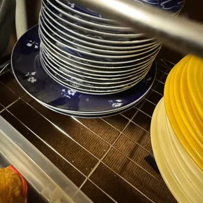 Sale Photo Thumbnail #117: Stacks of vintage ceramic plates, including a dark blue floral pattern and solid yellow and cream dinnerware, stored on a wire shelf.