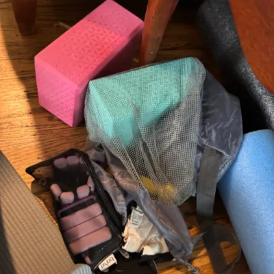 Sale Photo Thumbnail #295: Assorted yoga and fitness gear, including a pink foam yoga block, a teal block in a mesh bag, Bala bangles, and rolled mats on a hardwood floor.