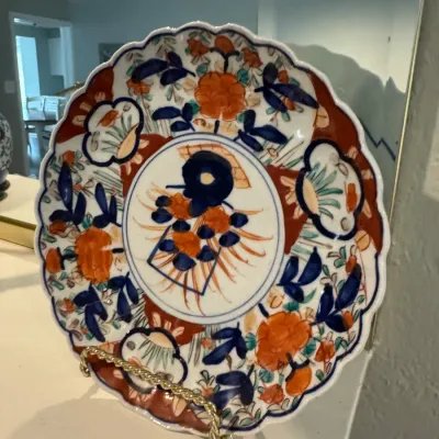 Sale Photo Thumbnail #59: Japanese Imari porcelain plate featuring a scalloped edge, hand-painted floral motifs in rust, cobalt blue, and gold, displayed on a brass wire stand.