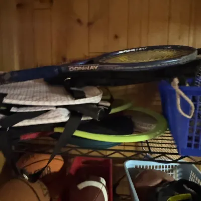 Sale Photo Thumbnail #150: Assorted sports equipment including a Donnay tennis racket, basketballs, and footballs stored on a wire shelving unit.