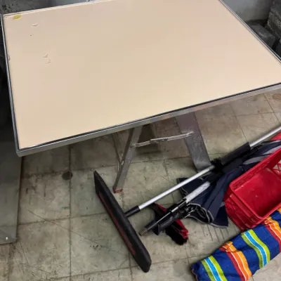 Sale Photo Thumbnail #96: Vintage folding card table with a beige vinyl top and metal frame, shown alongside various household items including a broom and a colorful striped bag.