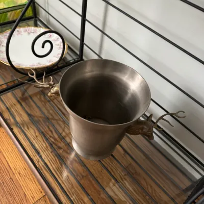 Sale Photo Thumbnail #328: Vintage pewter or stainless steel ice bucket featuring stag head handles with prominent antlers, displayed on a glass shelf.