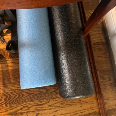 Sale Photo Thumbnail #296: Two foam rollers, one light blue and one black, stored on a hardwood floor beneath a wooden table.