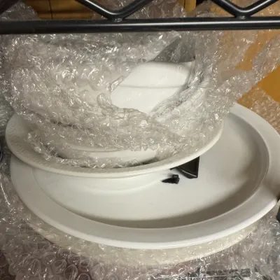 Sale Photo Thumbnail #155: Stack of white ceramic tableware, including plates and a serving dish, wrapped in protective bubble wrap on a wire shelf.