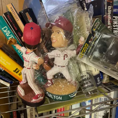 Sale Photo Thumbnail #16: Two Philadelphia Phillies bobbleheads, including a Steve Carlton figure in its original plastic packaging, displayed on a wire shelf with books and a 2004 World Series Champions shot glass.
