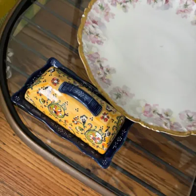Sale Photo Thumbnail #321: Vintage hand-painted ceramic butter dish with a vibrant yellow and blue floral pattern, shown alongside a delicate porcelain plate featuring pink floral details and a gold-gilded rim.