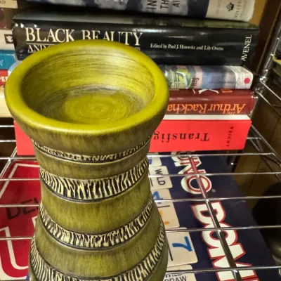 Sale Photo Thumbnail #119: Vintage olive green ceramic vase or candle holder with textured, zebra-like black banding, displayed on a wire shelf in front of books and board games.