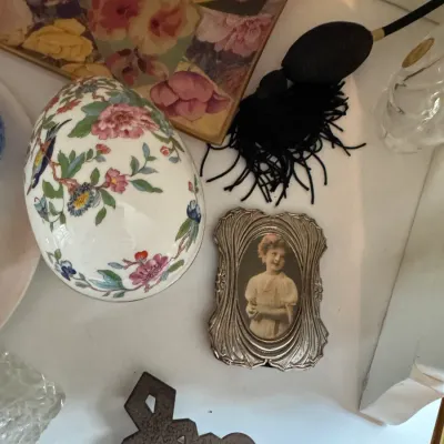 Sale Photo Thumbnail #179: Vintage vanity items including a floral porcelain egg trinket box, a small ornate silver-tone picture frame with a portrait, and a black perfume atomizer bulb.