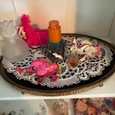 Sale Photo Thumbnail #181: Vintage vanity tray with brass trim and lace doily, displaying small perfume bottles and decorative fabric animal figurines, including a pink elephant and a lion.