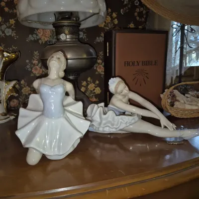 Sale Photo Thumbnail #9: Pair of vintage porcelain ballerina figurines displayed on a wooden table alongside an oil lamp and a Holy Bible.