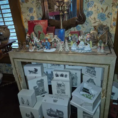 Sale Photo Thumbnail #33: Collection of Department 56 Dickens' Village collectibles displayed on and under a nailhead-trimmed console table, featuring numerous boxed ceramic buildings and figurine displays.