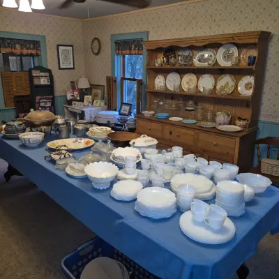 Sale Photo Thumbnail #280: Large collection of milk glass dishware displayed on a blue table, alongside a wooden hutch filled with decorative china plates and glassware.