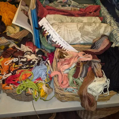 Sale Photo Thumbnail #80: Assorted craft and needlework supplies, including woven baskets filled with colorful yarn remnants, embroidery thread, fabric rolls, and a color chart.