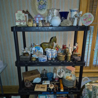 Sale Photo Thumbnail #94: Black plastic shelving unit holding an array of vintage collectibles, including figurines, glassware, porcelain trinket boxes, decorative plates, and books.