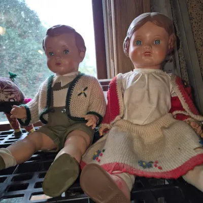Sale Photo Thumbnail #12: Pair of vintage mid-century composition or hard plastic dolls dressed in traditional European-style knitted outfits, sitting on a shelf.