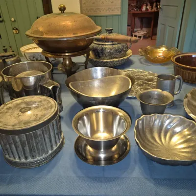 Sale Photo Thumbnail #273: Assortment of vintage metallic kitchenware and serveware, including a copper chafing dish, metal flour sifter, pewter cups, molded baking tins, and stoneware bowls displayed on a table.
