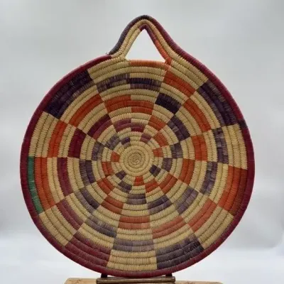 Sale Photo Thumbnail #16: Large hand-woven African Bolga basket platter featuring a colorful geometric pattern in orange, purple, and natural tones, complete with a built-in hanging loop.