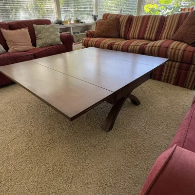 Large wooden square coffee table with pull-out extension leaves and an X-shaped trestle base, surrounded by upholstered sofas.