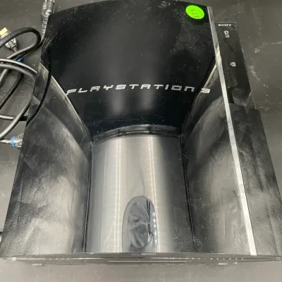 Original model Sony PlayStation 3 console in glossy black finish, showing some surface wear and dust, complete with power cord.