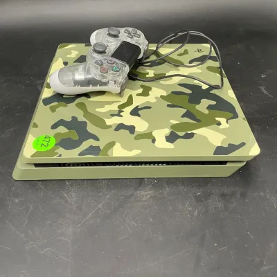 PlayStation 4 Slim console with a green camouflage skin, paired with a clear translucent DualShock 4 controller and charging cable.