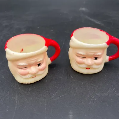 Pair of vintage winking Santa Claus miniature mugs, likely mid-century plastic or hard rubber, showing some paint wear consistent with age.