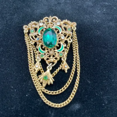 Vintage gold-tone filigree brooch featuring a large oval green rhinestone, green enamel accents, dangling charms, and draped curb chains.
