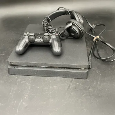 Sony PlayStation 4 Slim console in black, accompanied by a DualShock 4 wireless controller, wired over-ear headphones, and a power cable.