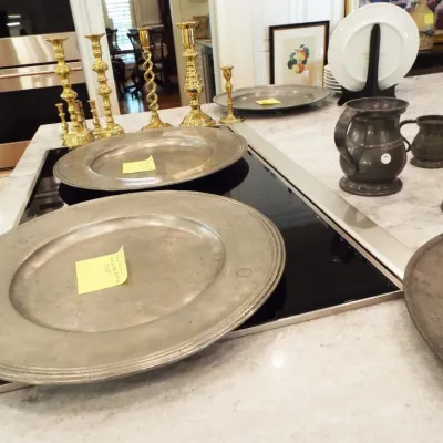 Collection of vintage pewter platters and tankards alongside polished brass candlesticks, arranged on a marble kitchen island.
