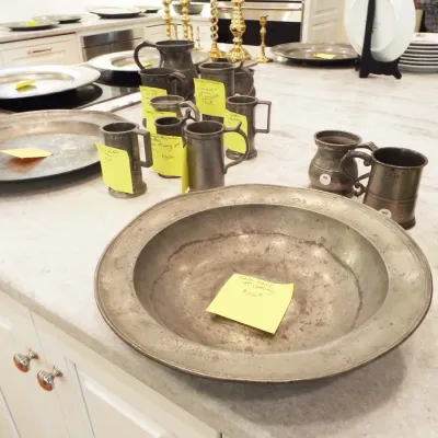 Collection of antique pewter serveware, including chargers, tankards, mugs, and a large deep bowl displayed on a countertop.