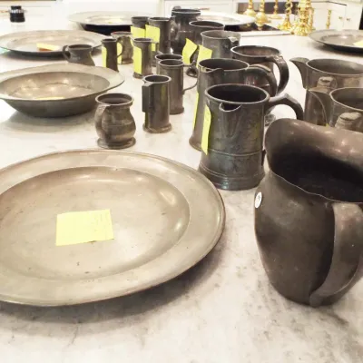 Large collection of antique pewter tableware, including platters, pitchers, tankards, and measure cups arranged on a marble countertop.
