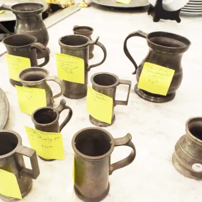Collection of antique pewter drinkware and pitchers, including various tankards and measures with handwritten descriptive tags.