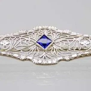 Art Deco style filigree bar pin featuring a central square-cut blue sapphire-colored gemstone set in white metal.