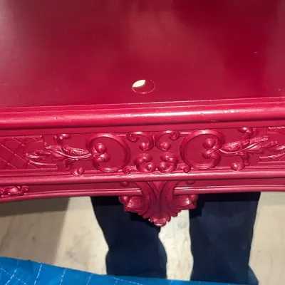 Sale Photo Thumbnail #78: Ornate wooden table painted in a deep burgundy red, featuring a small chip on the top surface and detailed scrollwork carving along the apron.