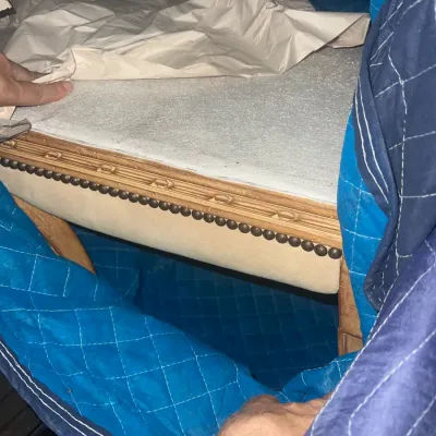 Sale Photo Thumbnail #89: Upholstered bench or stool featuring a light-colored fabric seat, carved wood trim, and brass nailhead detailing, partially wrapped in protective moving blankets.