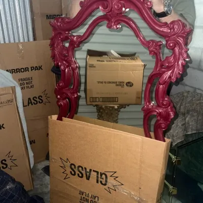 Sale Photo Thumbnail #60: Ornate Rococo-style mirror frame painted in a deep magenta or burgundy hue, shown partially packed in a cardboard mirror box.