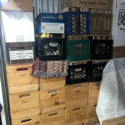 Sale Photo Thumbnail #12: Large collection of vinyl records stored in plastic milk crates and cardboard boxes, stacked alongside wooden storage crates in a storage unit.