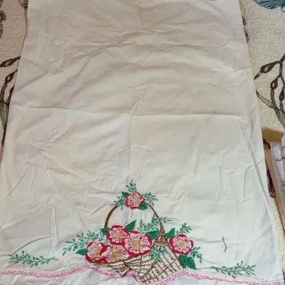 Vintage white cotton pillowcase featuring hand-embroidered pink flowers in a basket with a scalloped, pink crocheted edge.