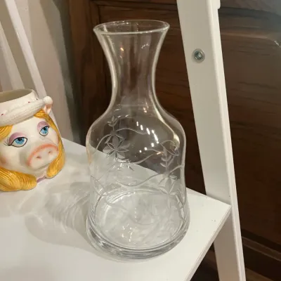 Vintage clear glass decanter or carafe featuring an etched floral and vine pattern, displayed alongside a Miss Piggy ceramic mug.