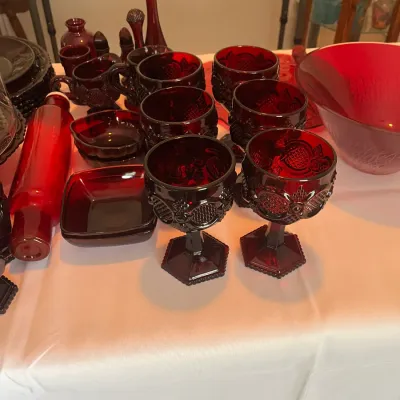 Collection of vintage ruby red glassware, including goblets, mugs, small bowls, and a rolling pin, displayed on a white tablecloth.