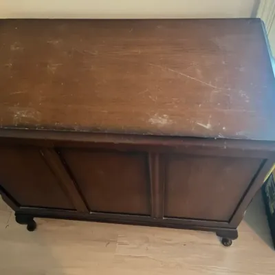 Vintage dark wood chest or trunk with paneled sides and short cabriole legs, showing significant surface wear and scratching on the top.