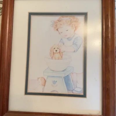 Framed pastel illustration of a young girl washing a puppy in a bowl, presented in a matted wooden frame.