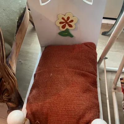 Vintage white wooden doll bed featuring a painted flower detail on the headboard and a textured orange cushion.