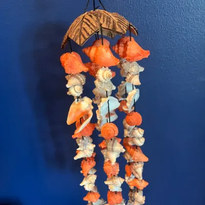 Handcrafted seashell wind chime featuring a coconut shell top and strands of orange and white shells, suspended against a blue wall.