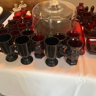 Collection of vintage ruby red glassware, including a covered cake stand, footed mugs, goblets, and small bowls.
