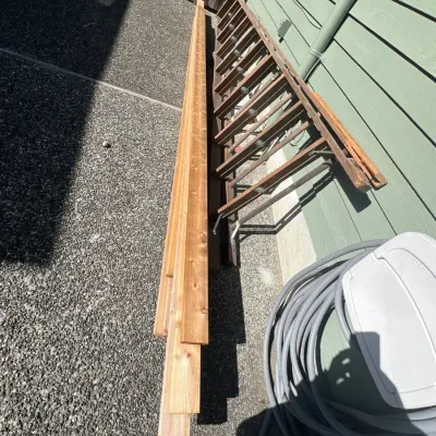 Stack of long lumber boards alongside vintage wooden extension ladders resting against a home's exterior siding.