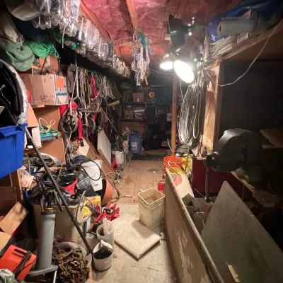 Crowded workshop passageway filled with tools, hardware, work lights, buckets, and stored supplies under exposed ceiling insulation.