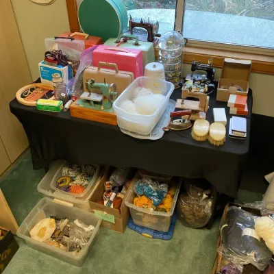 Collection of vintage sewing supplies, toy sewing machines, and train cases arranged on a table with bins of yarn below.
