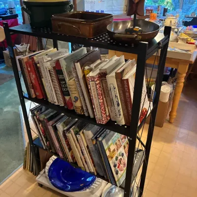 Black metal four-tier shelving unit filled with cookbooks, copper cookware, a slow cooker, and decorative glassware.